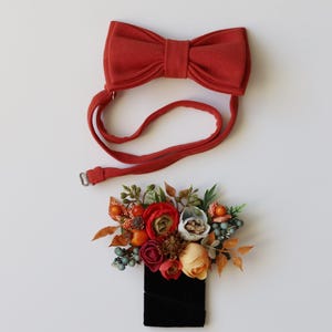 May include: A rust-colored fabric bow tie with an adjustable strap and a matching boutonniere featuring artificial flowers in shades of red, orange, and cream, accented with small green berries, brown pinecones, and foliage on a black base.