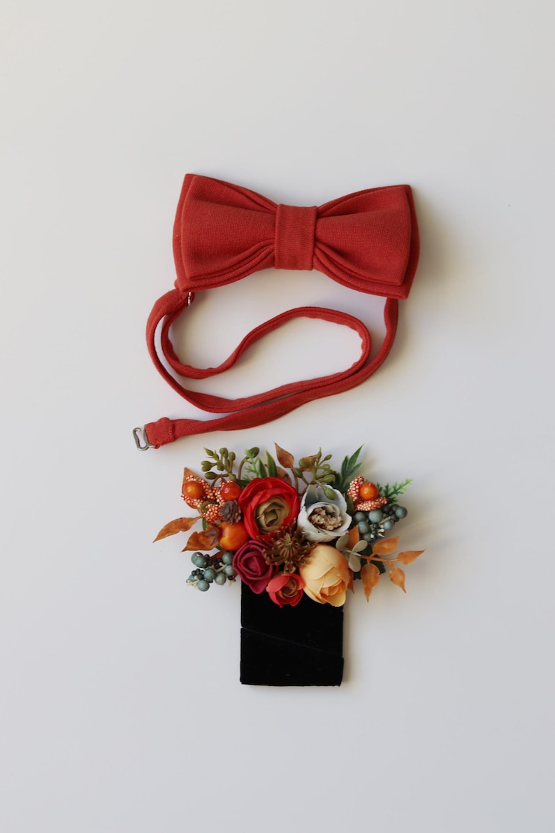 May include: A rust-colored fabric bow tie with an adjustable strap and a matching boutonniere featuring artificial flowers in shades of red, orange, and cream, accented with small green berries, brown pinecones, and foliage on a black base.