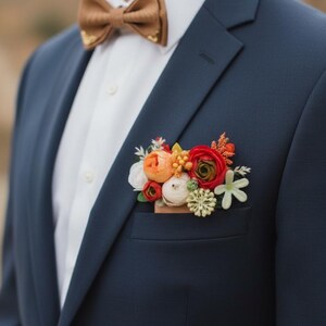 May include: A navy blue blazer worn with a white collared shirt and a tan bow tie. A floral pocket square accessory features a cluster of artificial flowers in shades of red, orange, white, and green tucked into the blazer's chest pocket.
