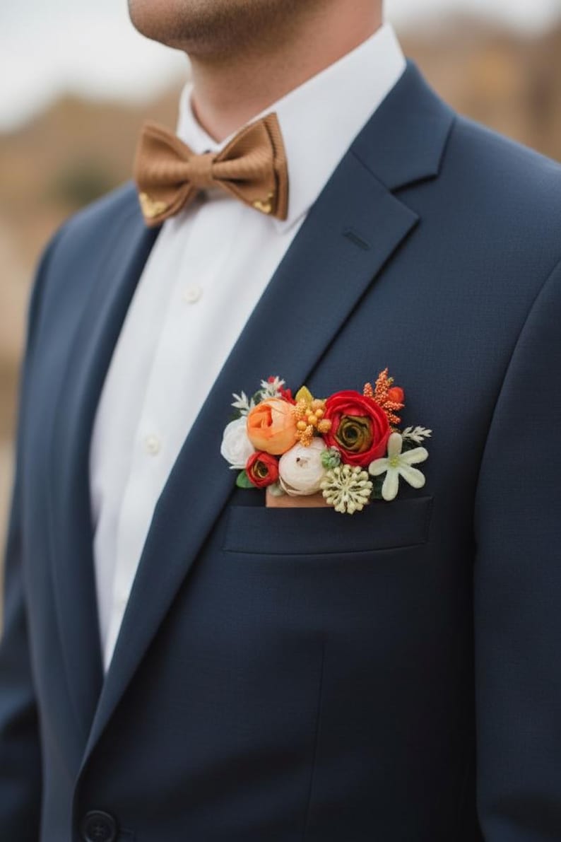 May include: A navy blue blazer worn with a white collared shirt and a tan bow tie. A floral pocket square accessory features a cluster of artificial flowers in shades of red, orange, white, and green tucked into the blazer's chest pocket.