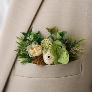May include: A beige blazer featuring a pocket square arrangement composed of artificial cream-colored roses, light green succulents, and various dark green foliage sprigs tucked neatly into the breast pocket of the textured fabric jacket.