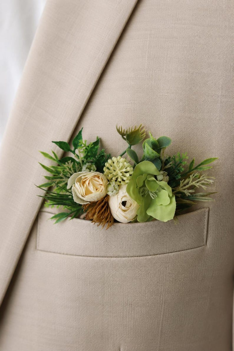 May include: A beige blazer featuring a pocket square arrangement composed of artificial cream-colored roses, light green succulents, and various dark green foliage sprigs tucked neatly into the breast pocket of the textured fabric jacket.