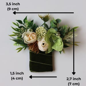 May include: A floral boutonniere featuring cream-colored ranunculus, green succulents, and assorted foliage wrapped in dark green velvet ribbon. The arrangement measures 3.5 inches wide by 2.7 inches high, with a 1.5-inch wide base.