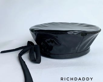 May include: A black patent leather beret with a glossy finish and a smooth, rounded top. The hat features a matching black fabric tie detail hanging from the side. The text "RICHDADDY BERET" is printed in black capital letters in the lower right.
