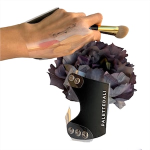 Può includere: Un porta palette per trucco nero con la scritta "PALETTEDALI" e uno scudo di plastica trasparente. Un pennello per trucco è tenuto sopra una mano con campioni di trucco. Un fiore viola scuro è sullo sfondo.