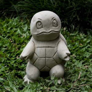 Op de afbeelding: Een grijs betonnen beeld van een schildpad met een gedetailleerde schild en grote ogen. Het beeld staat op een bed van groen gras.