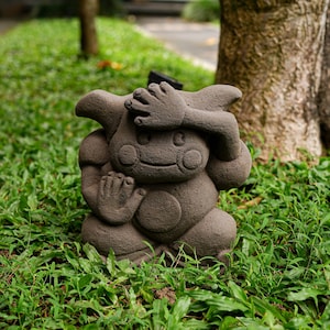 May include: A gray stone sculpture of a cartoon character with its hands covering its eyes. The sculpture is sitting on a bed of green grass.
