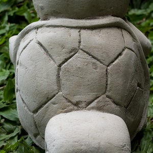 Pu&ograve; includere: Una statua di tartaruga in cemento grigio con un design a guscio dettagliato. La tartaruga &egrave; seduta su un letto di erba verde.