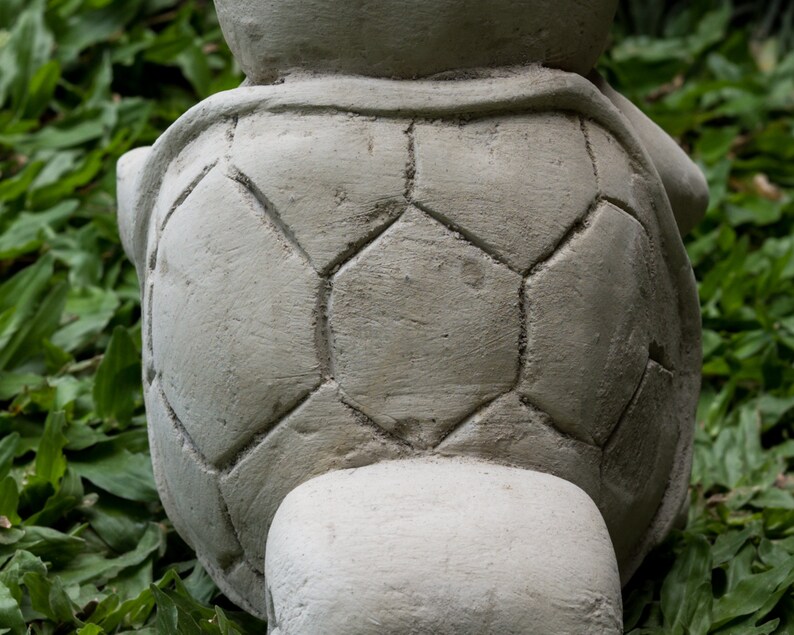 Pu&ograve; includere: Una statua di tartaruga in cemento grigio con un design a guscio dettagliato. La tartaruga &egrave; seduta su un letto di erba verde.