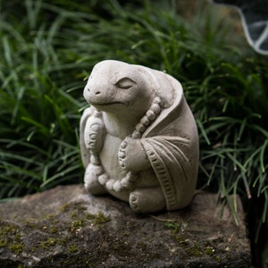 May include: A small, gray stone sculpture of a turtle wearing a robe and holding a string of beads. The turtle is sitting on a rock with green grass in the background.