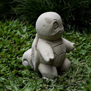 Pu&ograve; includere: Una scultura in cemento grigio di una tartaruga con un guscio, in piedi su un letto di erba verde.