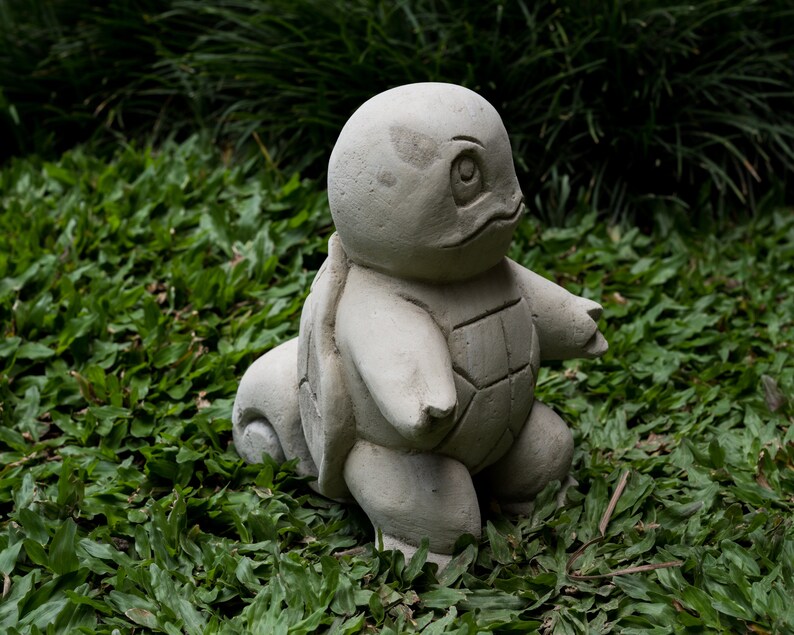 Pu&ograve; includere: Una scultura in cemento grigio di una tartaruga con un guscio, in piedi su un letto di erba verde.