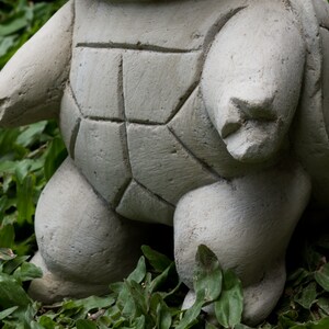 Pu&ograve; includere: Una statua di tartaruga in cemento grigio con un guscio dettagliato e una superficie testurizzata. La tartaruga &egrave; in piedi su un letto di erba verde.