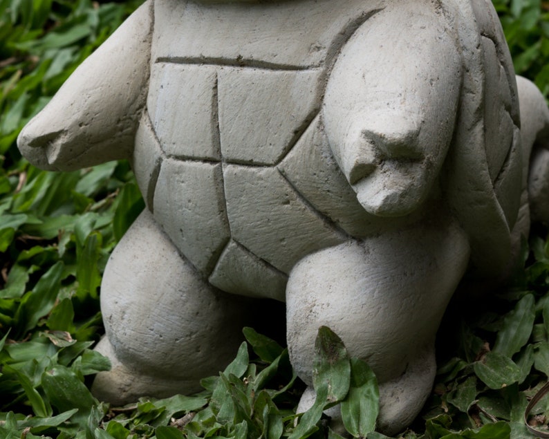 Pu&ograve; includere: Una statua di tartaruga in cemento grigio con un guscio dettagliato e una superficie testurizzata. La tartaruga &egrave; in piedi su un letto di erba verde.
