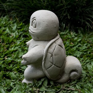 Pu&ograve; includere: Una scultura di tartaruga in cemento grigio con un guscio dettagliato, seduta su un letto di erba verde.