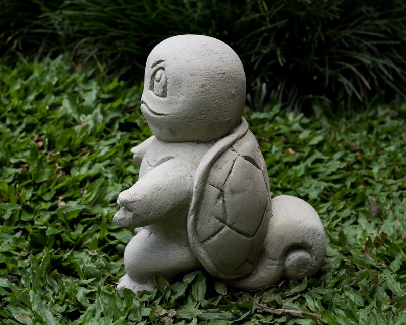 Pu&ograve; includere: Una scultura di tartaruga in cemento grigio con un guscio dettagliato, seduta su un letto di erba verde.