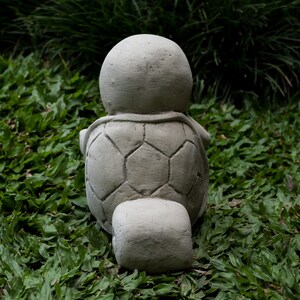 Pu&ograve; includere: Una statua di tartaruga in cemento grigio con un guscio dettagliato, seduta su un letto di erba verde.