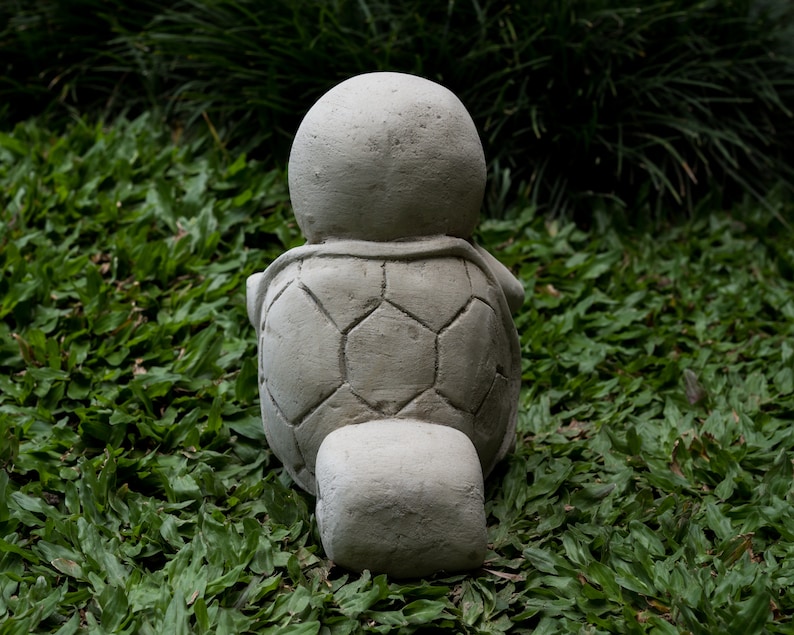 Pu&ograve; includere: Una statua di tartaruga in cemento grigio con un guscio dettagliato, seduta su un letto di erba verde.