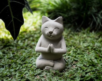 May include: A gray stone cat statue sitting in a meditative pose with its paws folded in front of its chest. The cat has a serene expression on its face.