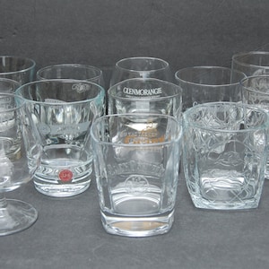 May include: A collection of clear glass tumblers with various etched logos, including Glenmorangie, Jack Daniel's, and Old No. 7. The glasses are arranged on a gray surface.