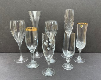 May include: A collection of eight clear glass champagne flutes with gold accents. Some of the flutes have etched designs.