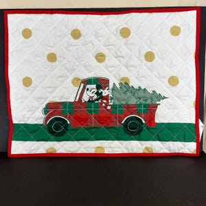 May include: A white quilted throw pillow with a red truck carrying a Christmas tree. Mickey Mouse and Minnie Mouse are in the truck. The truck has a red and black plaid design. The background is white with gold polka dots.