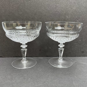 May include: Two clear glass champagne coupes with a diamond pattern on the bowl and stem. The coupes are on a gray surface.