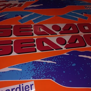 K&ouml;nnte beinhalten: Zwei rote und wei&szlig;e Aufkleber mit dem Text "Sea Doo" auf orangefarbenem Hintergrund. Die Aufkleber sind von blauen und wei&szlig;en abstrakten Designs umgeben.