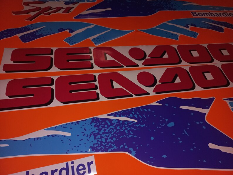 K&ouml;nnte beinhalten: Zwei rote und wei&szlig;e Aufkleber mit dem Text "Sea Doo" auf orangefarbenem Hintergrund. Die Aufkleber sind von blauen und wei&szlig;en abstrakten Designs umgeben.