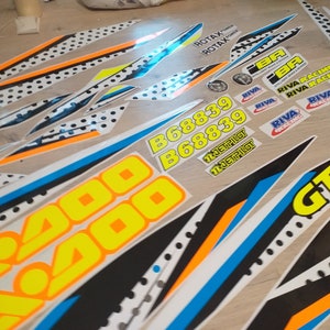 May include: A set of motorcycle decals featuring a black and white chequered pattern, yellow and orange accents, and the text "200", "B68839", "JETPILOT", "RIVA RACING", and "ROTAX POWER".