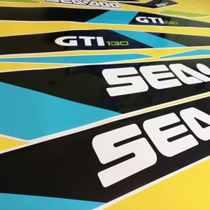 以下が含まれることがあります： 青色のアクセントが付いた白黒のシー-ドゥーのステッカーのクローズアップ。ステッカーには「シー-ドゥー」と「GTI 130」の文字が書かれています。