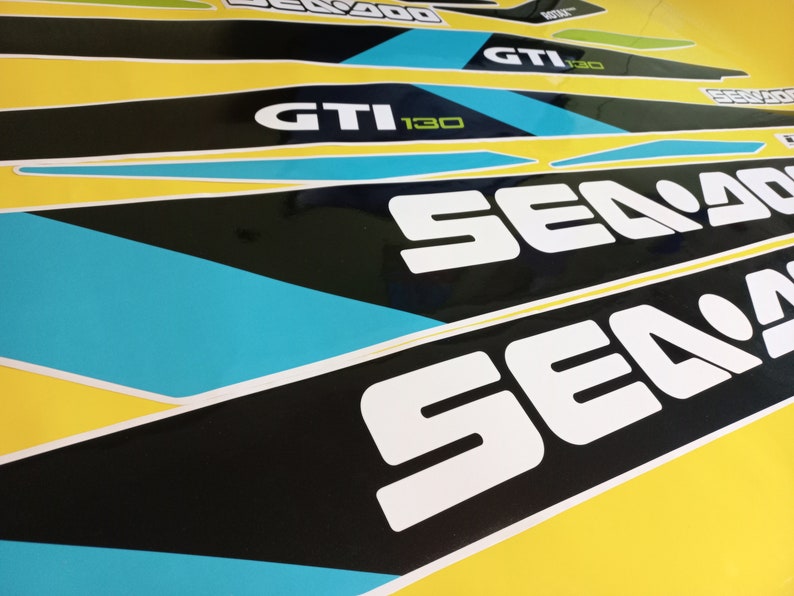 以下が含まれることがあります： 青色のアクセントが付いた白黒のシー-ドゥーのステッカーのクローズアップ。ステッカーには「シー-ドゥー」と「GTI 130」の文字が書かれています。