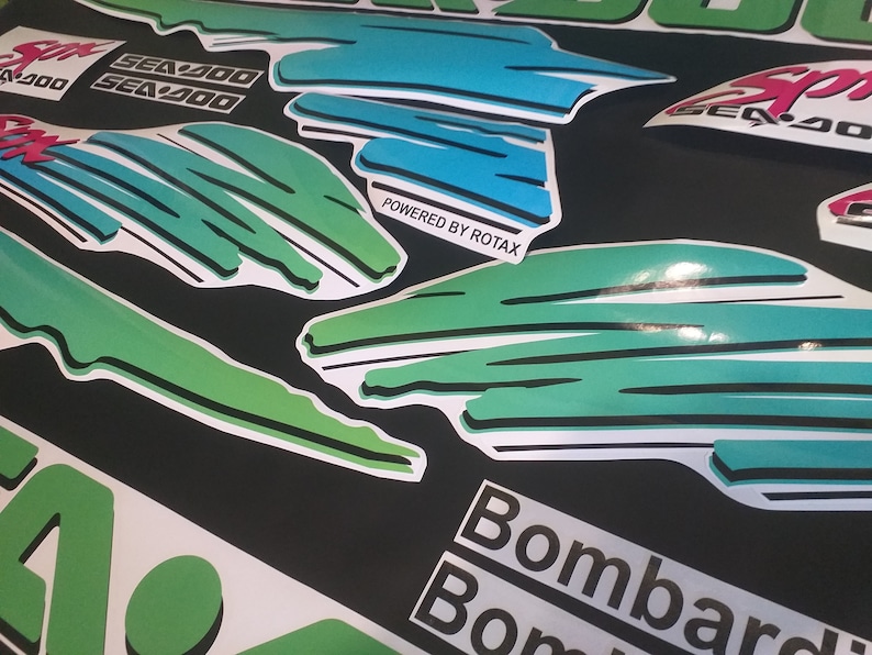 Puede incluir: Un conjunto de calcoman&iacute;as verdes, azules y blancas con el texto "Sea-Doo" y "Powered by Rotax". Las calcoman&iacute;as est&aacute;n dise&ntilde;adas para una embarcaci&oacute;n.