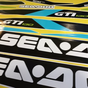 以下が含まれることがあります： 白黒のシー-ドゥーのデカールをクローズアップ。テキストは「Sea-Doo」と「GTI 130」で白。デカールは黄色の背景に貼られています。