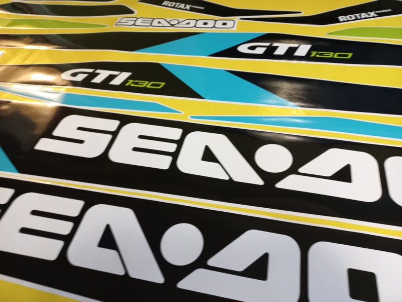 以下が含まれることがあります： 白黒のシー-ドゥーのデカールをクローズアップ。テキストは「Sea-Doo」と「GTI 130」で白。デカールは黄色の背景に貼られています。