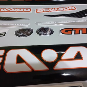 May include: A set of black and white decals with orange accents for a Sea-Doo watercraft. The decals include the text "Sea-Doo", "GTII 130", "Closed-Loop Cooling System", "Pop Cooling System", and "Rotax".