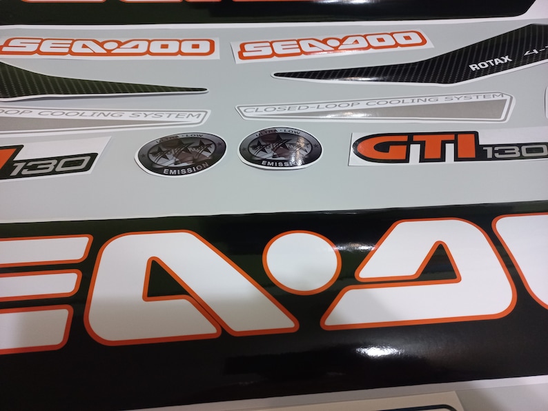 May include: A set of black and white decals with orange accents for a Sea-Doo watercraft. The decals include the text "Sea-Doo", "GTII 130", "Closed-Loop Cooling System", "Pop Cooling System", and "Rotax".