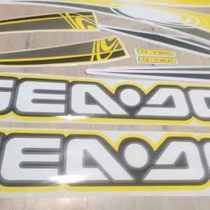 May include: Two white and gray "Sea-Doo" decals with a yellow outline. The decals are on a wooden surface.
