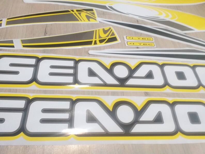 May include: Two white and gray "Sea-Doo" decals with a yellow outline. The decals are on a wooden surface.