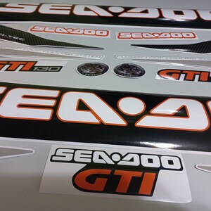 May include: A set of black and white Sea-Doo GTI 130 decals with orange accents. The decals feature the Sea-Doo logo, the model name, and the engine size. The decals are designed to be applied to a Sea-Doo watercraft.