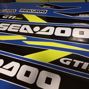May include: A set of black and white decals with yellow accents for a Sea-Doo GTI 130 watercraft. The decals feature the Sea-Doo logo and the text "GTI 130".