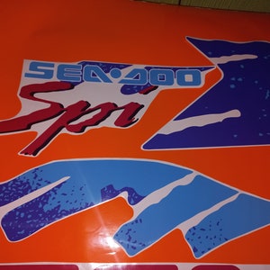 K&ouml;nnte beinhalten: Eine blaue und wei&szlig;e Grafik mit dem Text "SEA-DOO Sport" in Rot und Blau auf orangefarbenem Hintergrund.