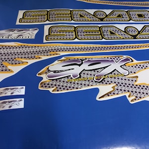 Puede incluir: Un conjunto de calcoman&iacute;as para moto acu&aacute;tica Sea-Doo blancas y doradas con un logotipo "Sea-Doo SPX Powered by Rotax Marine" negro y morado. Las calcoman&iacute;as presentan un patr&oacute;n de panal y un dise&ntilde;o de llama estilizado.