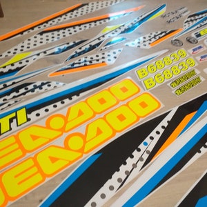May include: A set of motorcycle decals featuring a black, white, blue and orange colour scheme. The decals include the words "Sea Doo" and "GTI" in yellow lettering. The decals also include the numbers "668839" in yellow lettering.