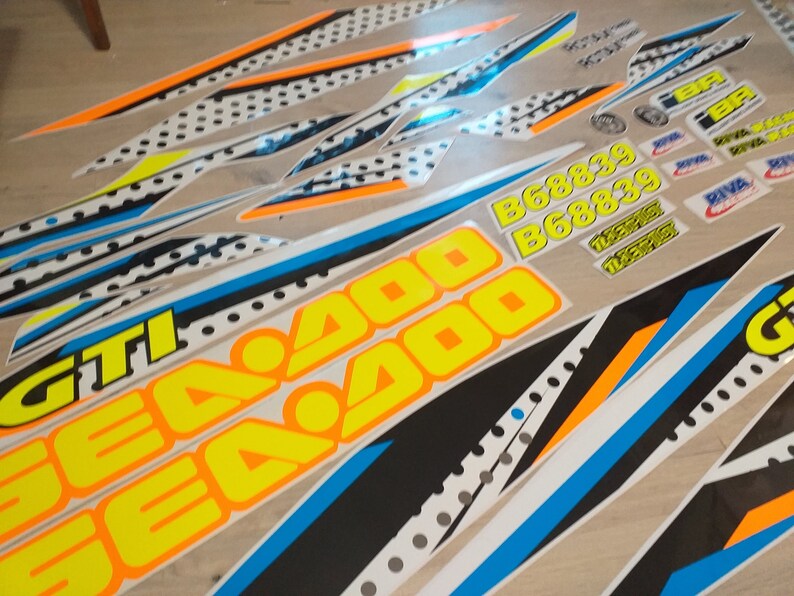 May include: A set of motorcycle decals featuring a black, white, blue and orange colour scheme. The decals include the words "Sea Doo" and "GTI" in yellow lettering. The decals also include the numbers "668839" in yellow lettering.