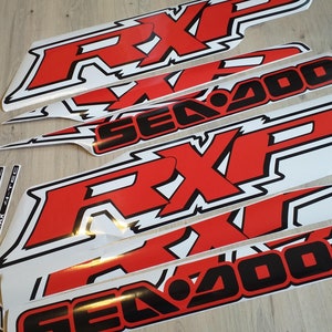 May include: A set of red and black vinyl decals for a Sea-Doo watercraft. The decals feature the words "Sea-Doo" and "RXP" in a bold, stylized font. The decals are designed to be applied to the hull of a Sea-Doo watercraft.