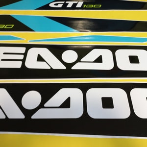 以下が含まれることがあります： 黄色と黒の背景に、白黒のシー-ドゥーのロゴが映し出されています。ロゴは「Sea-Doo」という文字が白で書かれており、「Sea」の文字の真ん中に白い円があります。