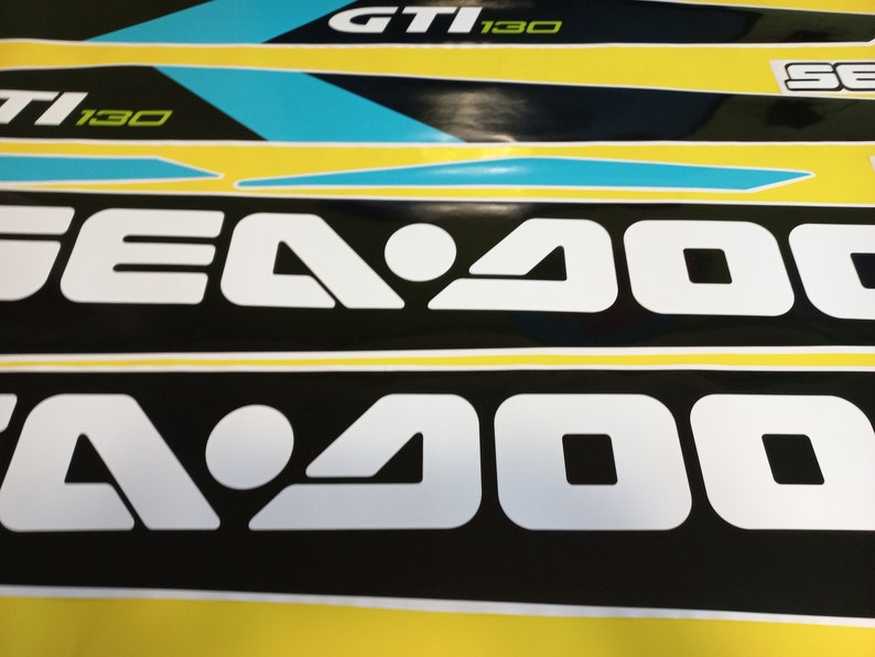 以下が含まれることがあります： 黄色と黒の背景に、白黒のシー-ドゥーのロゴが映し出されています。ロゴは「Sea-Doo」という文字が白で書かれており、「Sea」の文字の真ん中に白い円があります。