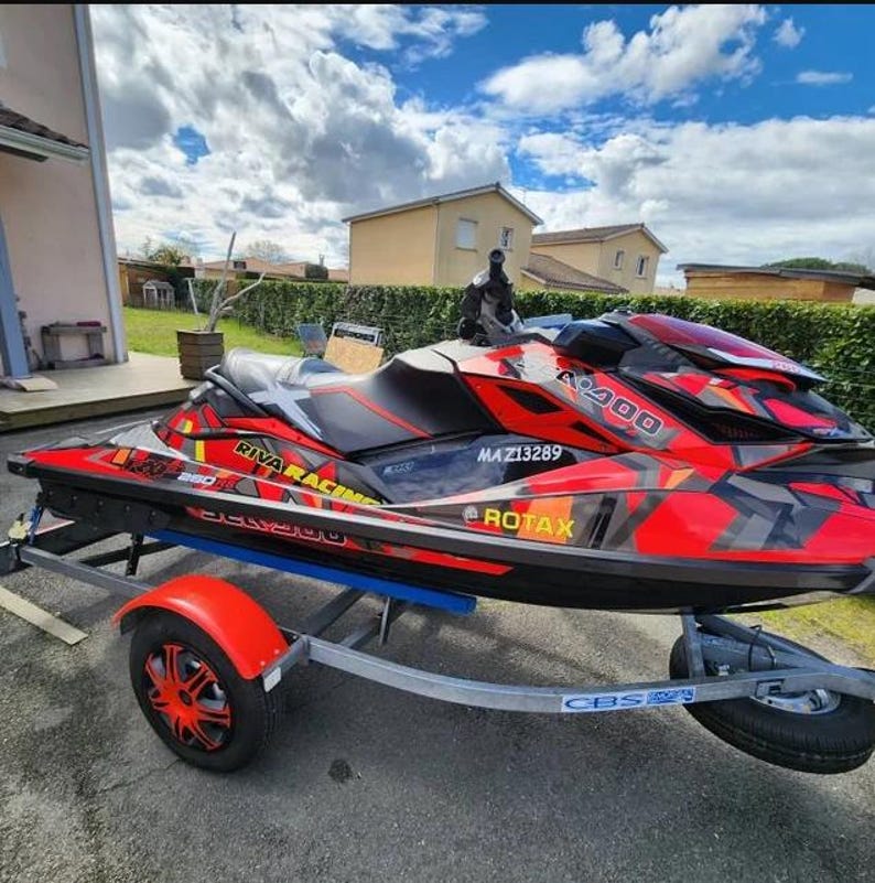 Puede incluir: Una moto de agua roja, negra y gris con un logotipo "RIVA RACING" negro y amarillo y un logotipo "ROTAX" negro y amarillo. La moto de agua est&aacute; en un remolque con un logotipo "CBS" negro y blanco.