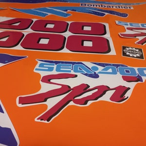 K&ouml;nnte beinhalten: Ein Satz roter und blauer Sea-Doo-Aufkleber mit dem Text "Sea-Doo" und "Sea-Doo Sport" sowie der Nummer "2000". Die Aufkleber befinden sich auf einem leuchtend orangenen Hintergrund.